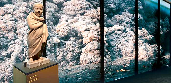 Skulptur eines Knaben im Mantel vor der Fotografie einer Pyroklastischen Wolke